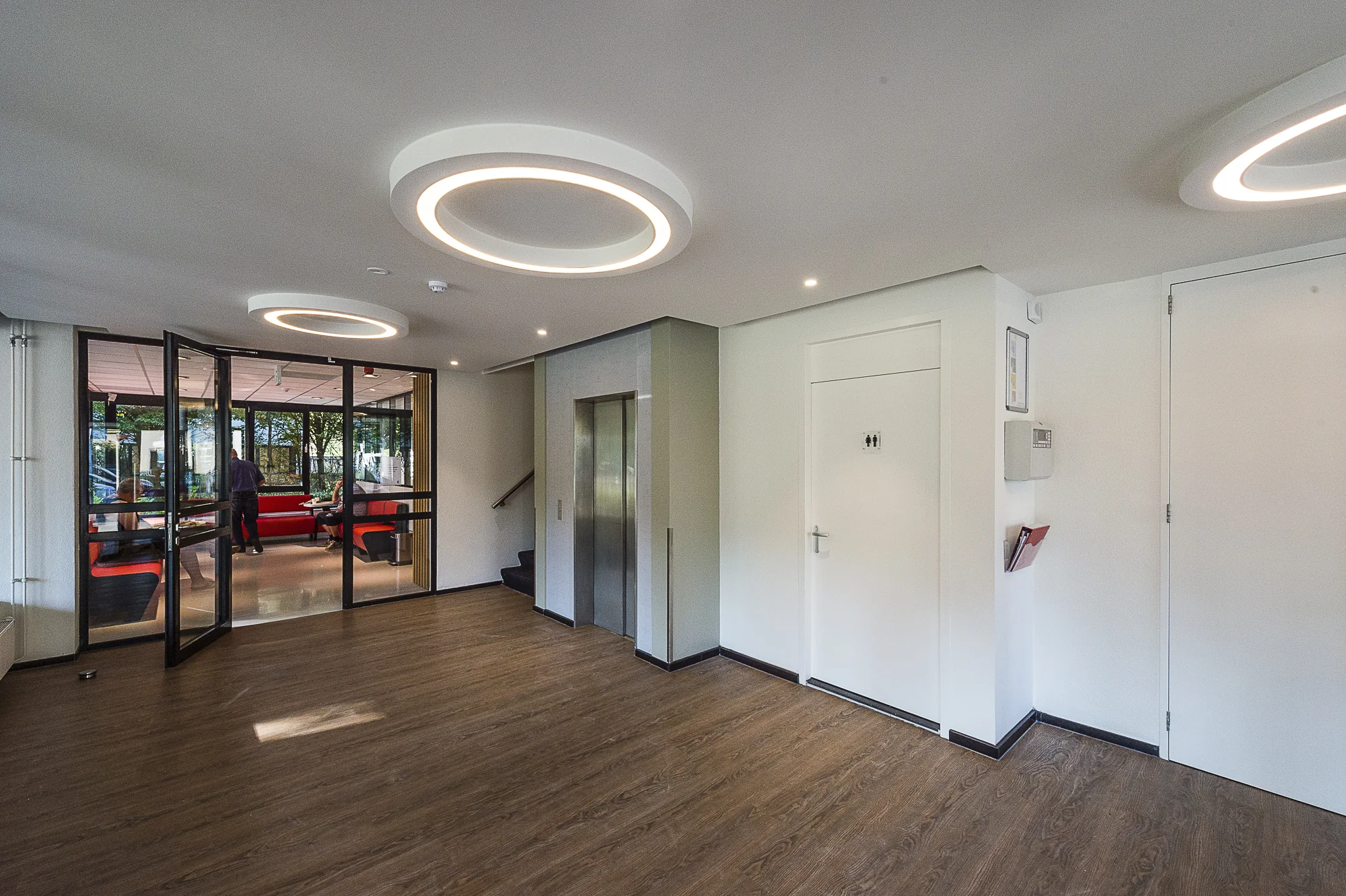 Moderne entreehal aan de Max Euwelaan met houten vloer, ronde plafondlampen, lift en wachtruimte achter glazen deuren.